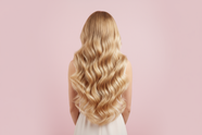 A women showing her behind very lucious and thick hair that are blond. She is standing in the middle of the photo. The hair are the vocal point, she is blond herself. The background is pink, but very light pink