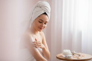 An image of a woman taking care of her body, putting cream on her body having a towel on her hair. The background is pink, but very light pink